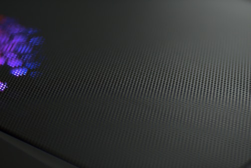 A close-up view of a perforated metal surface with a reflective texture. Subtle purple and blue light glimmers on one side, creating a dynamic contrast against the dark, matte finish of the surface.