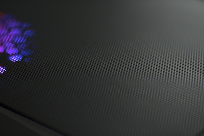A close-up view of a perforated metal surface with a reflective texture. Subtle purple and blue light glimmers on one side, creating a dynamic contrast against the dark, matte finish of the surface.