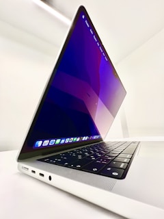 A sleek, modern laptop with a backlit keyboard and a thin profile is positioned at an angle. The screen displays an abstract purple and blue wallpaper, indicating a powered-on state. Multiple application icons are visible on the dock. The laptop is set against a minimalist white background, emphasizing its elegant design.