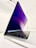 A sleek, modern laptop with a backlit keyboard and a thin profile is positioned at an angle. The screen displays an abstract purple and blue wallpaper, indicating a powered-on state. Multiple application icons are visible on the dock. The laptop is set against a minimalist white background, emphasizing its elegant design.