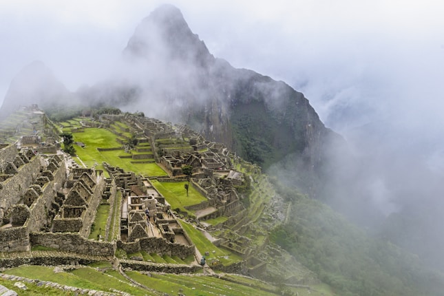 Ancient stone ruins are nestled within lush green terraces high in the mountains, partially shrouded by misty clouds. The structures are intricately arranged, showcasing terraces and pathways.