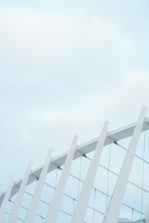 White, modern architectural structure featuring several parallel beams connected by horizontal wires, set against a pale blue sky with soft clouds.