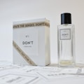 A minimalist design featuring a perfume bottle and its packaging. The bottle is transparent with a simple label, while the box is white with a bold font. A ribbon with the text 