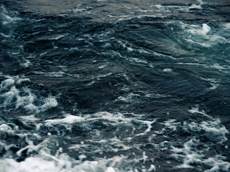 Dark ocean waves exhibit a dynamic and turbulent surface with white foam cresting over the deep, shadowy water.