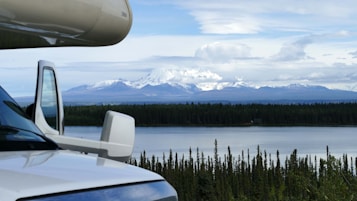 A scenic view depicts a snow-capped mountain range under a partly cloudy sky, with a lake and dense forest in the foreground. The side of a recreational vehicle is partially visible on the left, suggesting a travel or adventure theme.
