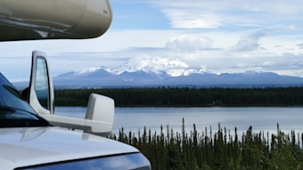 A scenic view depicts a snow-capped mountain range under a partly cloudy sky, with a lake and dense forest in the foreground. The side of a recreational vehicle is partially visible on the left, suggesting a travel or adventure theme.