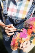 A person in a multicolored checkered shirt and denim overalls is holding a paint palette and mixing vibrant colors with a brush. The palette is filled with various hues like pink, orange, yellow, and green.