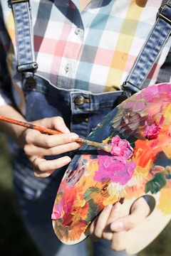 A person in a multicolored checkered shirt and denim overalls is holding a paint palette and mixing vibrant colors with a brush. The palette is filled with various hues like pink, orange, yellow, and green.