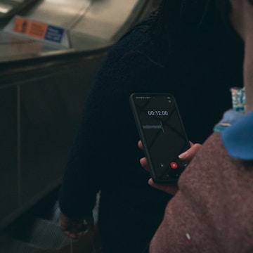 A person holding a smartphone while standing on an escalator, possibly recording audio or video. The phone screen is visible, displaying a timer and waveform. The scene is dimly lit, contributing to a somewhat muted atmosphere.