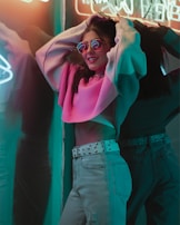 A person wearing pink-tinted sunglasses and a pink sweater poses confidently against a reflective surface. The setting is illuminated with colorful neon lights, creating a vibrant and lively atmosphere. The person's casual jeans and studded belt add a trendy touch to the overall look.
