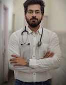 A man with a beard and glasses is standing confidently with his arms crossed. He is wearing a white button-up shirt and has a stethoscope around his neck, suggesting he is a medical professional. The background is a blurred indoor corridor.