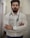 A man with a beard and glasses is standing confidently with his arms crossed. He is wearing a white button-up shirt and has a stethoscope around his neck, suggesting he is a medical professional. The background is a blurred indoor corridor.