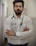 A man with a beard and glasses is standing confidently with his arms crossed. He is wearing a white button-up shirt and has a stethoscope around his neck, suggesting he is a medical professional. The background is a blurred indoor corridor.