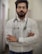 A man with a beard and glasses is standing confidently with his arms crossed. He is wearing a white button-up shirt and has a stethoscope around his neck, suggesting he is a medical professional. The background is a blurred indoor corridor.