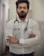A man with a beard and glasses is standing confidently with his arms crossed. He is wearing a white button-up shirt and has a stethoscope around his neck, suggesting he is a medical professional. The background is a blurred indoor corridor.