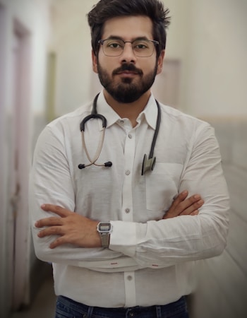 A man with a beard and glasses is standing confidently with his arms crossed. He is wearing a white button-up shirt and has a stethoscope around his neck, suggesting he is a medical professional. The background is a blurred indoor corridor.