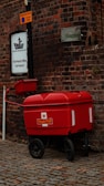 A bright red Royal Mail trolley is parked on a cobblestone street beside a brick building. The wall features a sign for Ship Safe Training Group Ltd. and a 'Restricted Zone' notice on a pole. The color of the trolley contrasts sharply with the dark, textured bricks.