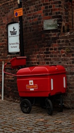 A bright red Royal Mail trolley is parked on a cobblestone street beside a brick building. The wall features a sign for Ship Safe Training Group Ltd. and a 'Restricted Zone' notice on a pole. The color of the trolley contrasts sharply with the dark, textured bricks.