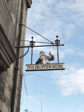 A hanging sign attached to the side of a building features a painted figure holding up a rolled document. Below the figure, the sign reads 'THE PEOPLE'S STORY'. The building appears to be made of stone, and the background shows a clear blue sky with scattered clouds.