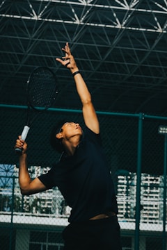 A person is poised to serve a tennis ball, holding a racket high above their head and looking upwards. The setting appears to be an indoor tennis court with a large, netted ceiling overhead. The lighting casts dramatic shadows, emphasizing the athlete's form and movement.
