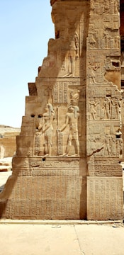 A large, intricately carved stone wall featuring Egyptian hieroglyphics and depictions of ancient Egyptian figures. The carvings show standing figures in traditional headdresses and garments, surrounded by columns of hieroglyphs. Small pieces of the stone wall seem worn and weathered, indicating age.