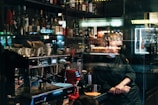 A dimly lit bar interior with a variety of liquor bottles on shelves. A barista stands behind a coffee machine, glancing to the side while wearing a mask under his chin. Cups are arranged on the counter, and reflections from the window create an intriguing visual effect.