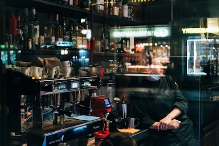 A dimly lit bar interior with a variety of liquor bottles on shelves. A barista stands behind a coffee machine, glancing to the side while wearing a mask under his chin. Cups are arranged on the counter, and reflections from the window create an intriguing visual effect.