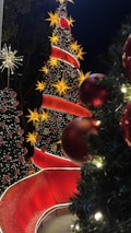 A festive scene featuring several Christmas trees adorned with bright lights and ornaments. The largest tree is decorated with large yellow star-shaped lights and a winding red ribbon, accompanied by red baubles. Other smaller trees complement the scene, surrounded by lush greenery and sparkling lights. The overall atmosphere is vibrant and celebratory, typical of holiday decorations.