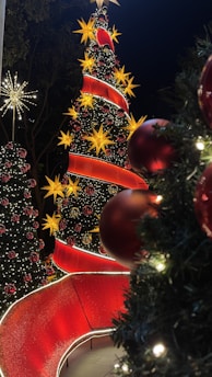 A festive scene featuring several Christmas trees adorned with bright lights and ornaments. The largest tree is decorated with large yellow star-shaped lights and a winding red ribbon, accompanied by red baubles. Other smaller trees complement the scene, surrounded by lush greenery and sparkling lights. The overall atmosphere is vibrant and celebratory, typical of holiday decorations.