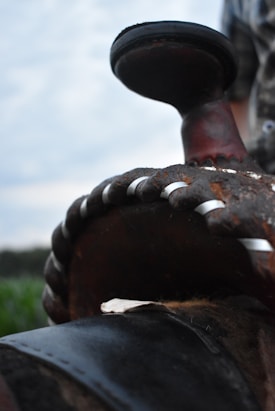 A close-up view of a worn-out saddle horn, showing detailed stitching and wear on the leather. The background is slightly blurred, depicting an outdoor scene.