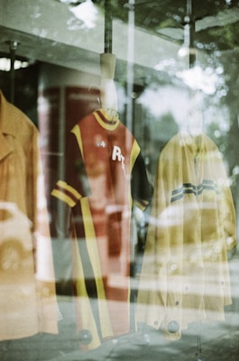 A display window featuring mannequins with vintage-style clothing. The garments include a red dress with yellow and black stripes and a yellow coat. Reflections from the glass and surrounding environment are visible, creating a layered visual effect.