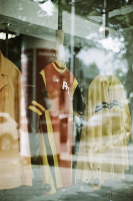 A display window featuring mannequins with vintage-style clothing. The garments include a red dress with yellow and black stripes and a yellow coat. Reflections from the glass and surrounding environment are visible, creating a layered visual effect.