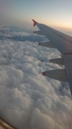An airplane wing with red-tipped winglets is visible, set against a backdrop of thick, fluffy clouds below. The sky above transitions from blue to a warm gradient, indicating either sunrise or sunset.