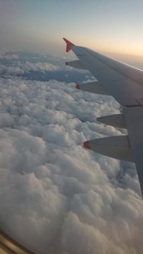 An airplane wing with red-tipped winglets is visible, set against a backdrop of thick, fluffy clouds below. The sky above transitions from blue to a warm gradient, indicating either sunrise or sunset.