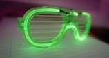 A pair of green plastic shutter sunglasses lies on a lined piece of paper. The glasses have a translucent, vibrant appearance, with horizontal slats instead of traditional lenses. The lighting gives the image a soft, ambient glow, with a background that fades into shades of purple and pink.