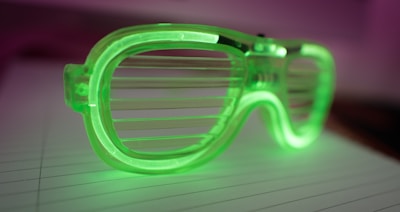 A pair of green plastic shutter sunglasses lies on a lined piece of paper. The glasses have a translucent, vibrant appearance, with horizontal slats instead of traditional lenses. The lighting gives the image a soft, ambient glow, with a background that fades into shades of purple and pink.