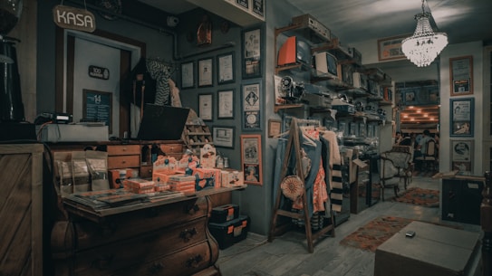A vintage store interior filled with retro items and decor. The left side features a wooden chest with merchandise and papers on top. The back wall has numerous framed certificates and posters, alongside vintage TV sets and other appliances on shelves. Clothes are displayed on a wooden rack, and there are chairs and tables in the background leading to a dining area. The room is lit by a chandelier, creating a nostalgic atmosphere.