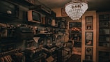A dimly lit room filled with vintage electronics and furnishings. Old televisions and radios are neatly arranged on shelves alongside vintage typewriters. An ornate chandelier hangs from the ceiling, casting a warm glow throughout the room. Framed posters and records adorn the walls, contributing to an antique and nostalgic atmosphere.