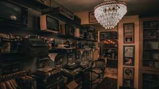 A dimly lit room filled with vintage electronics and furnishings. Old televisions and radios are neatly arranged on shelves alongside vintage typewriters. An ornate chandelier hangs from the ceiling, casting a warm glow throughout the room. Framed posters and records adorn the walls, contributing to an antique and nostalgic atmosphere.