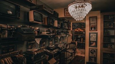 A dimly lit room filled with vintage electronics and furnishings. Old televisions and radios are neatly arranged on shelves alongside vintage typewriters. An ornate chandelier hangs from the ceiling, casting a warm glow throughout the room. Framed posters and records adorn the walls, contributing to an antique and nostalgic atmosphere.