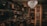 A dimly lit room filled with vintage electronics and furnishings. Old televisions and radios are neatly arranged on shelves alongside vintage typewriters. An ornate chandelier hangs from the ceiling, casting a warm glow throughout the room. Framed posters and records adorn the walls, contributing to an antique and nostalgic atmosphere.
