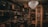 A dimly lit room filled with vintage electronics and furnishings. Old televisions and radios are neatly arranged on shelves alongside vintage typewriters. An ornate chandelier hangs from the ceiling, casting a warm glow throughout the room. Framed posters and records adorn the walls, contributing to an antique and nostalgic atmosphere.