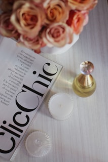 A tabletop displays a magazine with bold typography, surrounded by a bouquet of peach and pink roses in a white vase. Nearby, a glass perfume bottle with a golden cap is positioned alongside a lit white candle in a decorative holder. The setting conveys a sense of elegance and sophistication.