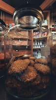 A large glass jar filled with round, sesame-coated cookies. The jar is placed on a shelf in a cozy market setting with wooden interiors. Various packaged snacks are visible in the background, adding a warm ambiance.