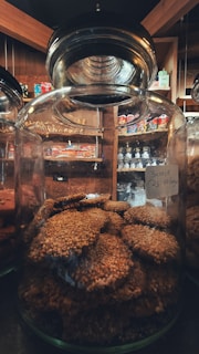 A large glass jar filled with round, sesame-coated cookies. The jar is placed on a shelf in a cozy market setting with wooden interiors. Various packaged snacks are visible in the background, adding a warm ambiance.