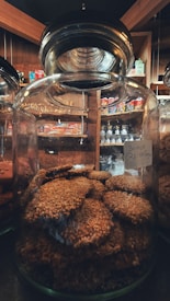 A large glass jar filled with round, sesame-coated cookies. The jar is placed on a shelf in a cozy market setting with wooden interiors. Various packaged snacks are visible in the background, adding a warm ambiance.