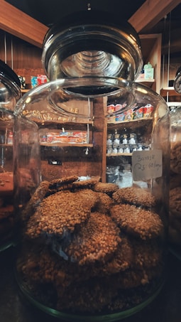 A large glass jar filled with round, sesame-coated cookies. The jar is placed on a shelf in a cozy market setting with wooden interiors. Various packaged snacks are visible in the background, adding a warm ambiance.