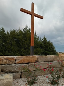 A large wooden cross stands prominently on a stone platform in front of a backdrop of dense evergreen trees. The sky is overcast, casting a muted light over the scene. In the foreground, small bushes with vibrant red flowers grow among the rocks.