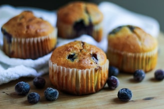 Four blueberry muffins are placed on a wooden surface surrounded by scattered blueberries. The muffins are golden brown with visible blueberries and have a paper lining. A soft focus in the background highlights a white cloth.