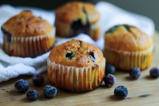 Four blueberry muffins are placed on a wooden surface surrounded by scattered blueberries. The muffins are golden brown with visible blueberries and have a paper lining. A soft focus in the background highlights a white cloth.
